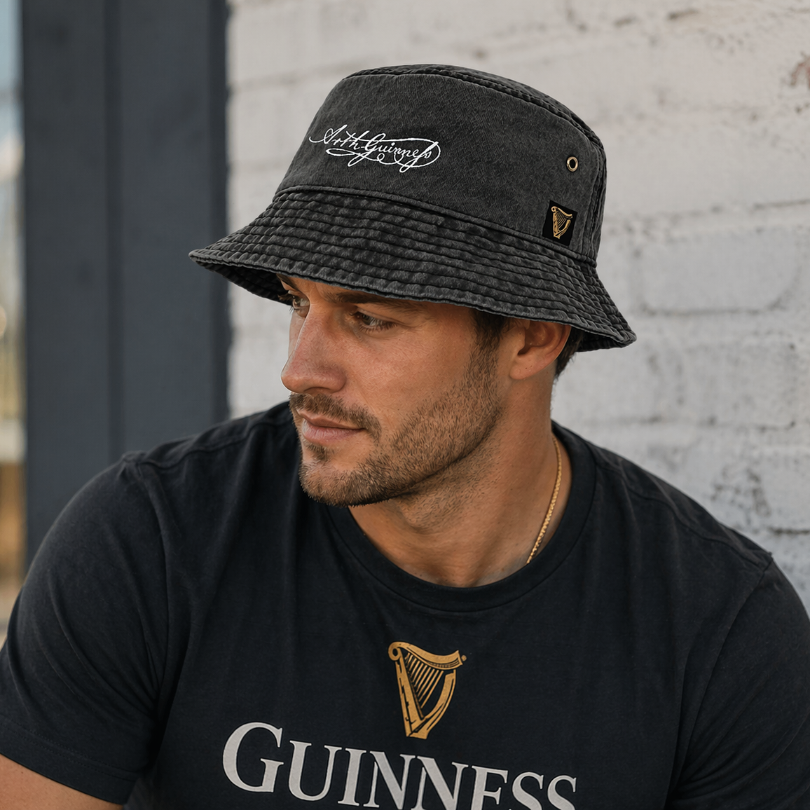 A man wearing a Guinness Signature Bucket Hat and a matching Guinness t-shirt sits in front of a white brick wall, looking to the side.