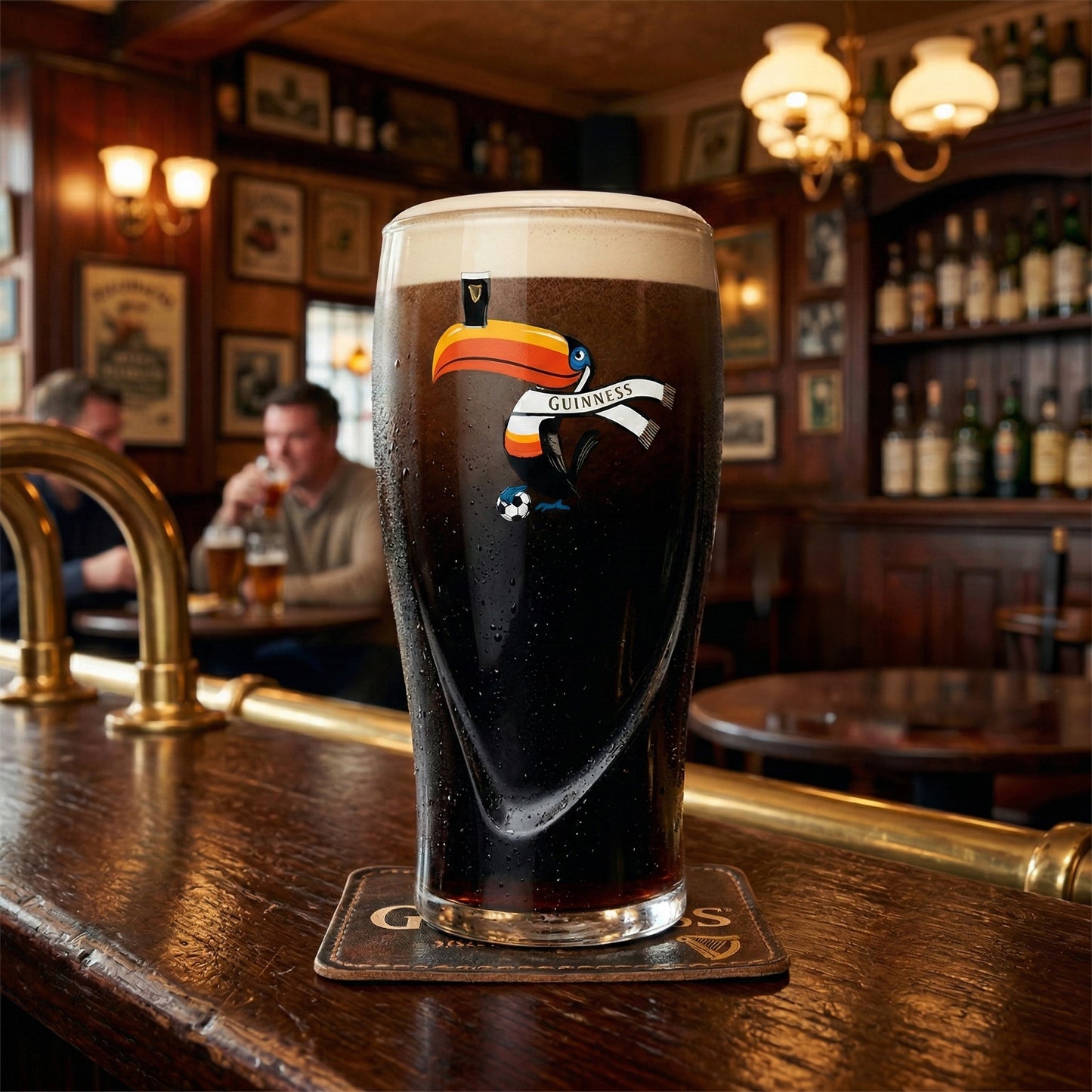 A pint of Guinness stout is poured into a Guinness Soccer Pint Glass – Special Edition from Guinness Webstore US, resting on a bar counter in a classic pub as two people chat in the blurred background.