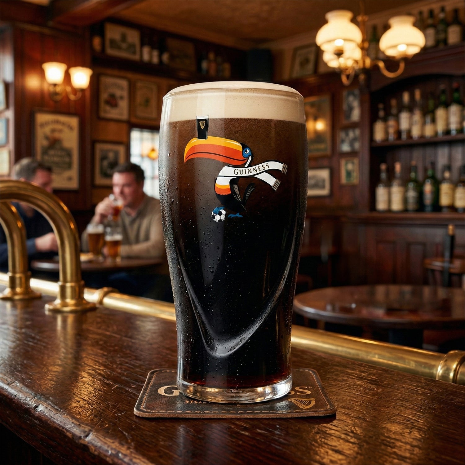 A pint of Guinness stout is poured into a Guinness Soccer Pint Glass – Special Edition from Guinness Webstore US, resting on a bar counter in a classic pub as two people chat in the blurred background.