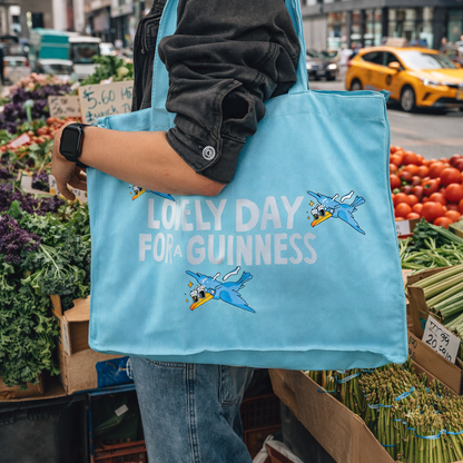 At an outdoor market, someone carries the blue Guinness Summer Collection Tote Bag from Guinness Webstore US, featuring cartoon planes and "Lovely Day For A Guinness"—the perfect oversized cotton accessory for summer shopping.