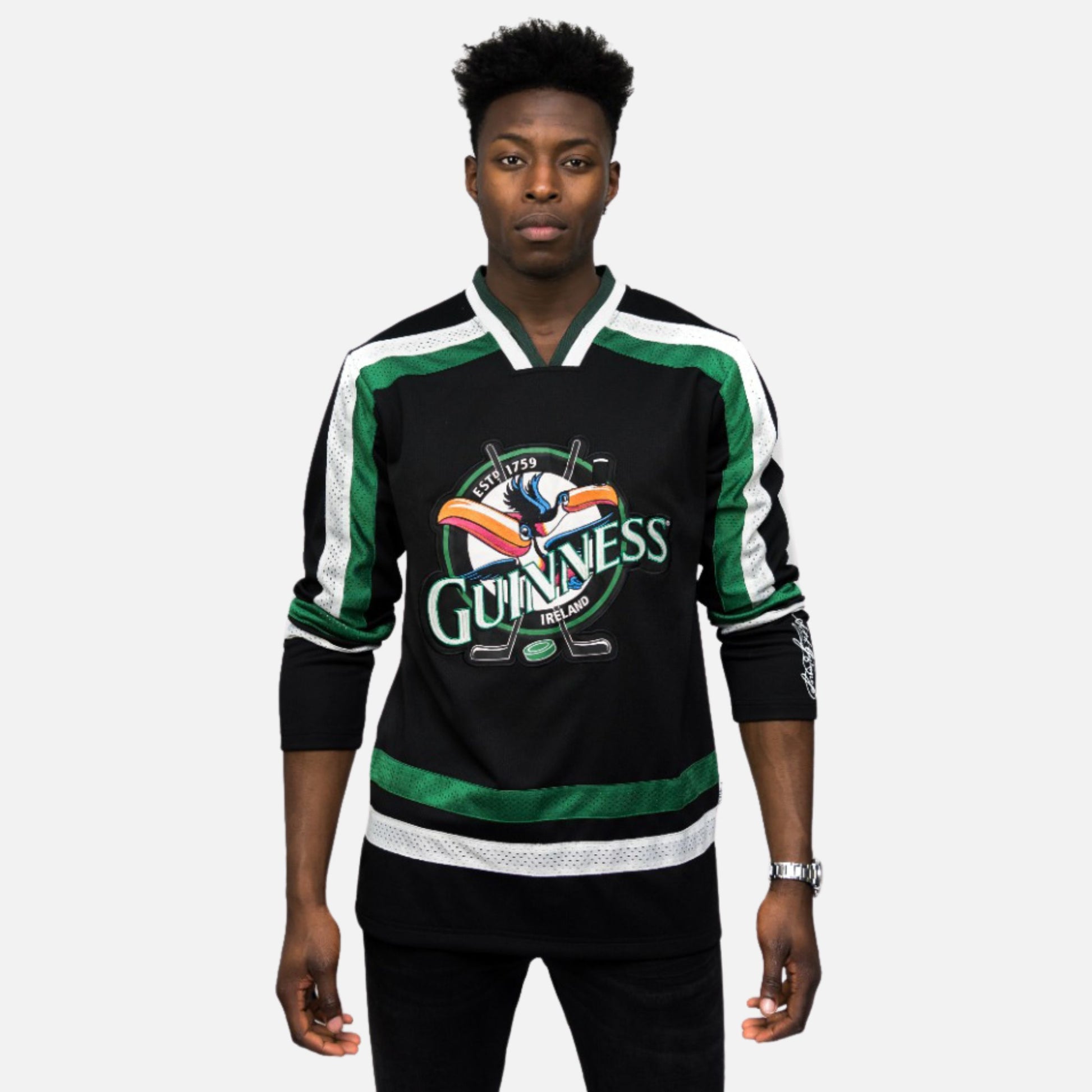 A man stands facing forward wearing the Guinness Toucan Hockey Jersey in black and green by Guinness, featuring white accents and a large Guinness logo with the iconic toucan on the front.