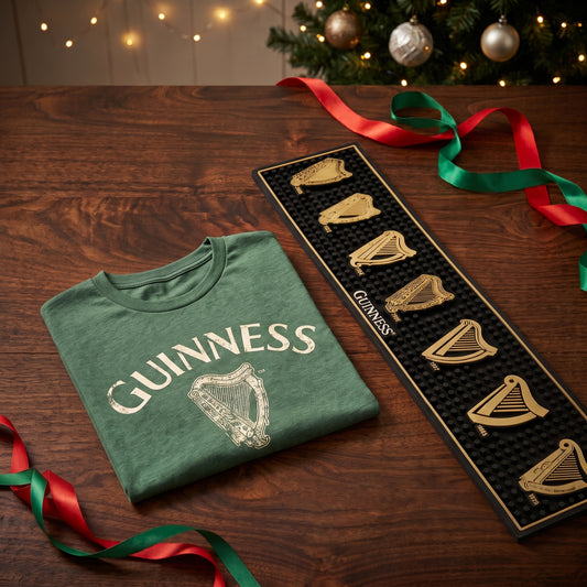 The Guinness Vintage Holiday Gift Set features a vintage green T-shirt with a gold harp logo and a black bar mat with six gold harp designs on a wooden table.