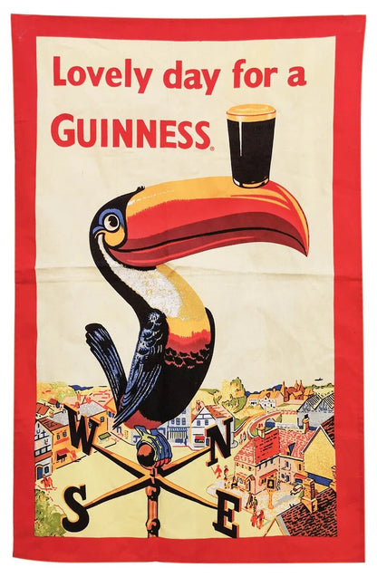 The Guinness Toucan Tea Towel by Guinness US features a whimsical illustration of a toucan balancing a pint of Guinness on its beak, accompanied by the phrase "Lovely day for a Guinness." The design is set against a vibrant backdrop of houses and weather vane elements, adding kitchen charm to any space.