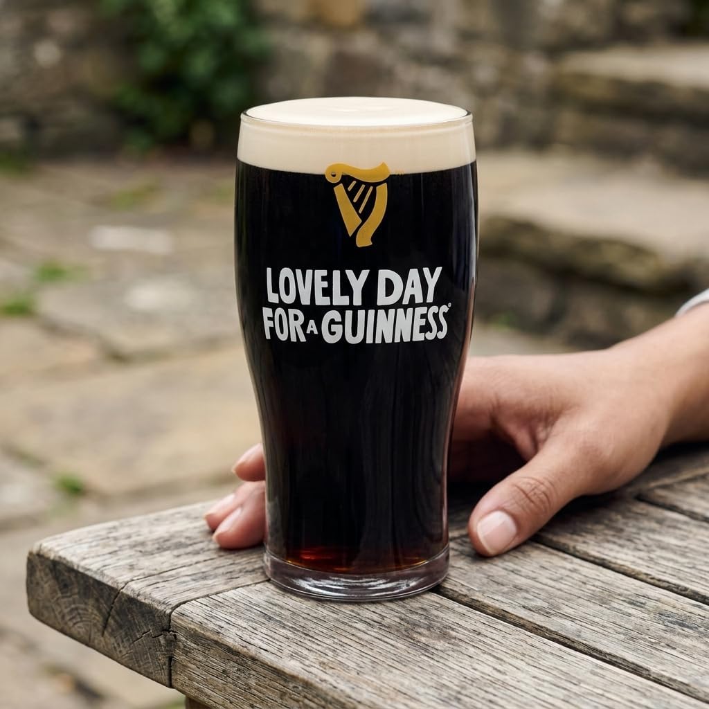 A hand rests on a wooden table beside the Guinness Summer Collection Lovely Day Pint Glass from Guinness Webstore US. "Lovely Day for a Guinness" is featured, with stone steps and outdoor scenery in the background.