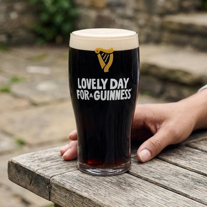 A hand rests on a wooden table beside the Guinness Summer Collection Lovely Day Pint Glass from Guinness Webstore US. "Lovely Day for a Guinness" is featured, with stone steps and outdoor scenery in the background.