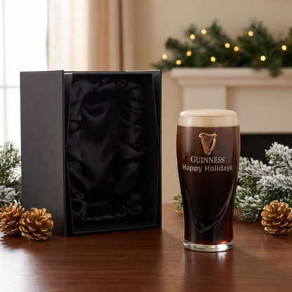 A Personalized Guinness Glass Gift Set from Guinness Webstore US sits by an open gift box, pine cones, greenery, and glowing holiday lights in the background.