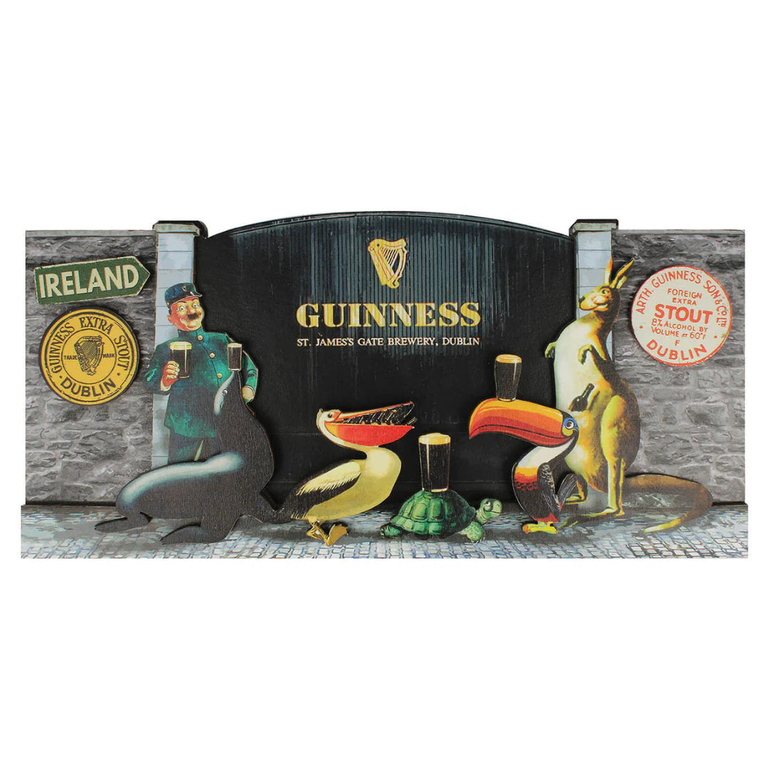 A cutout display inspired by John Gilroy artwork features a zookeeper and animals holding or admiring pints of Guinness in front of a brewery gate, with prominent Guinness branding, Irish stout signage, and the Guinness Gilroy Wooden Sign – 3D St James Gate from the Guinness Webstore US.