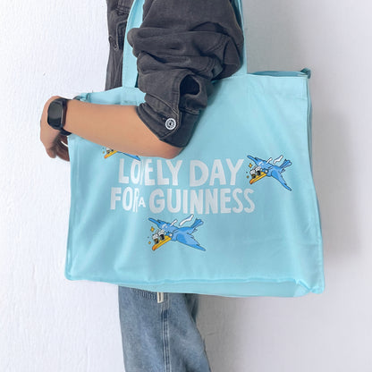 A person in a black jacket and jeans carries the Guinness Seb Curi Tote Bag - Blue from Guinness Webstore US, featuring cartoon birds and “LOVELY DAY FOR A GUINNESS,” in a style inspired by Sebastian Curi.