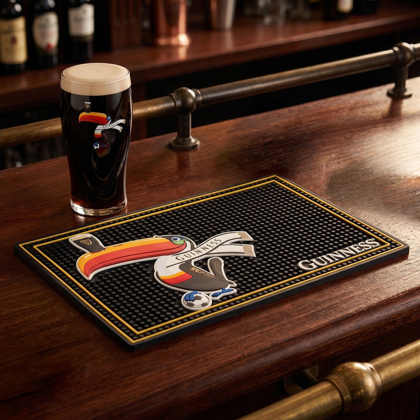 A pint of dark beer sits on a bar in the Guinness Soccer Matchday Pack from the Guinness Webstore US, featuring a soccer pint glass and a branded bar mat with a stylized toucan illustration.