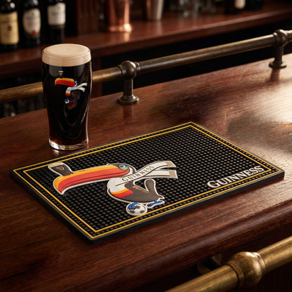 A pint of dark beer sits on a bar in the Guinness Soccer Matchday Pack from the Guinness Webstore US, featuring a soccer pint glass and a branded bar mat with a stylized toucan illustration.