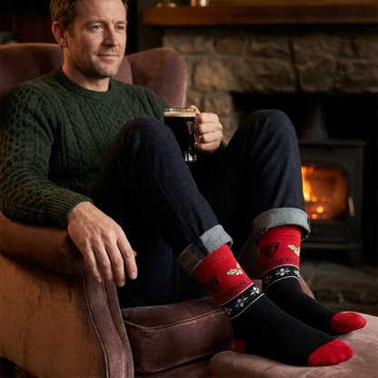 A man sitting in an armchair by a fireplace wears Guinness Harp & Holly Holiday Socks featuring a harp logo, holly leaves, and snowflakes.