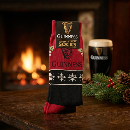Guinness Harp & Holly Holiday Socks in red and black with snowflake patterns and holly leaves, displayed on a wooden table next to a pint of stout and pine branches.