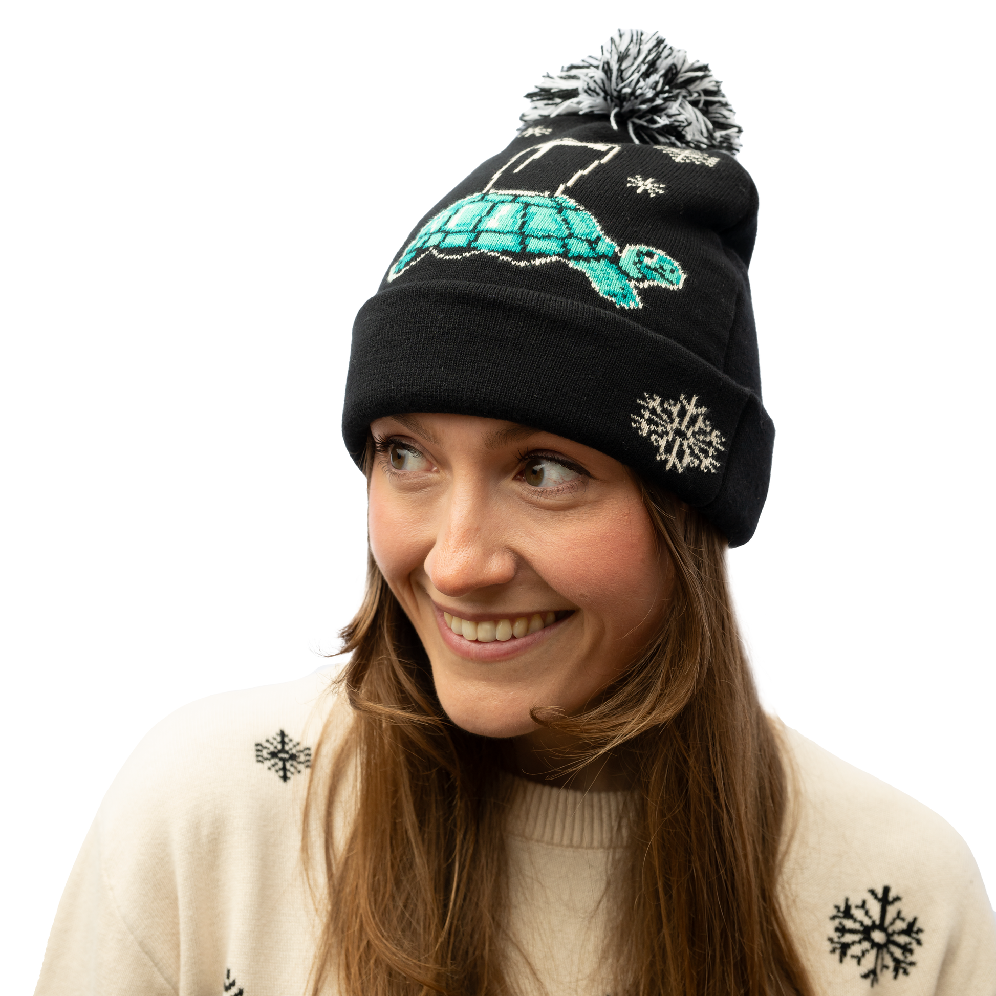 A woman with long hair is wearing a Guinness Holiday Tortoise Beanie from the Guinness Webstore US, which features a black design with a tortoise and snowflakes. She pairs it with a white sweater embellished with snowflakes, smiling as she looks to the side.