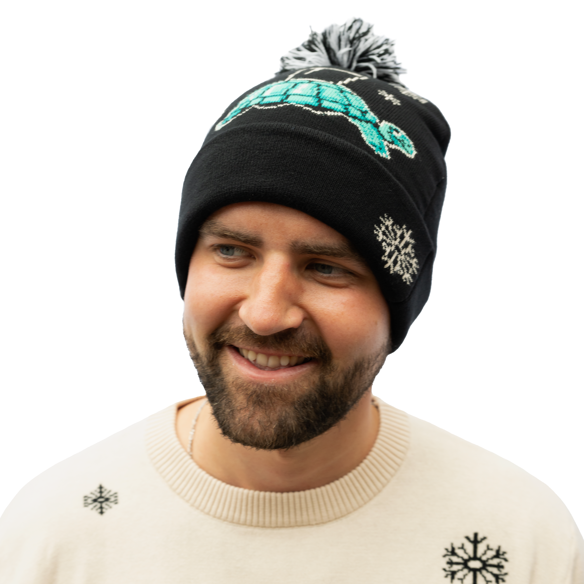 A man wearing the Guinness Holiday Tortoise Beanie from the Guinness Webstore US, featuring a black beanie with a tortoise motif and snowflake design, smiles warmly in his cream sweater adorned with snowflakes, showcasing his winter closet favorites.