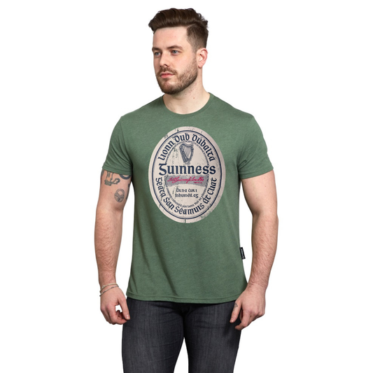 A man with short brown hair and a beard wears the Guinness Green Distressed Label Tee, a cotton blend unisex graphic t-shirt with a circular logo, standing against a plain white background.