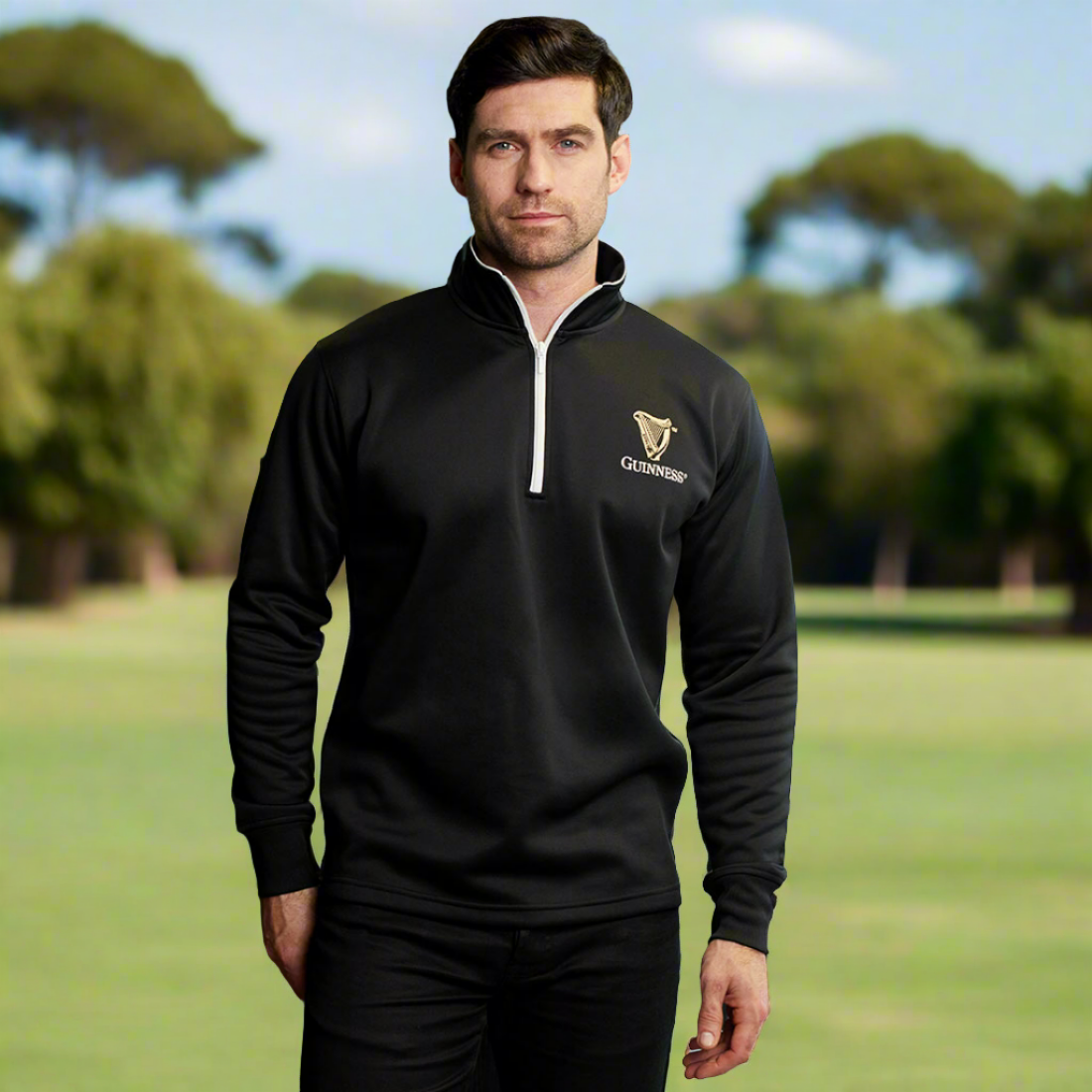 A man stands outdoors on grass, wearing a black Guinness Harp Half Zip Sweater with moisture-wicking properties. Trees are visible in the blurred background.