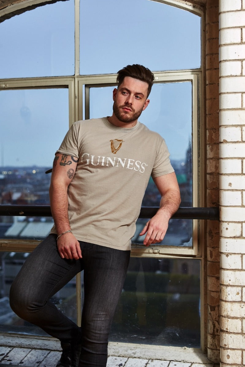 A man wearing a Guinness Trademark Label T-Shirt Beige leans against a window.
