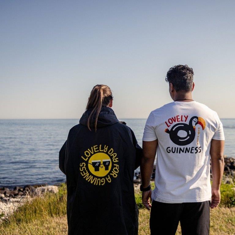 Two people enjoying a summer day by the ocean in their Guinness "Lovely Day for a Guinness" Toucan White Tee t-shirts.