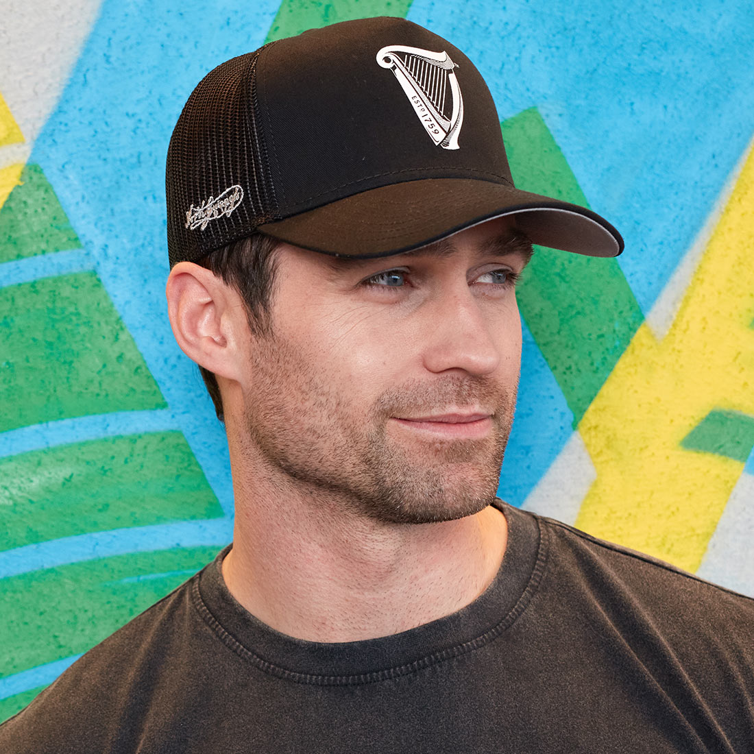 A man wearing a black hat with the Guinness Premium Black & White Cap logo in front of a colorful wall.