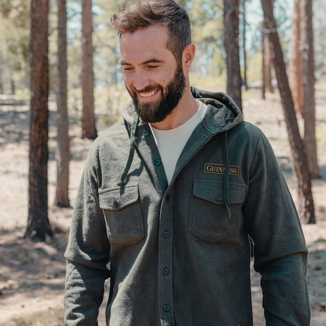 A man with a beard smiling in the woods while wearing a Guinness Deep Green Shirt Jacket hoodie.