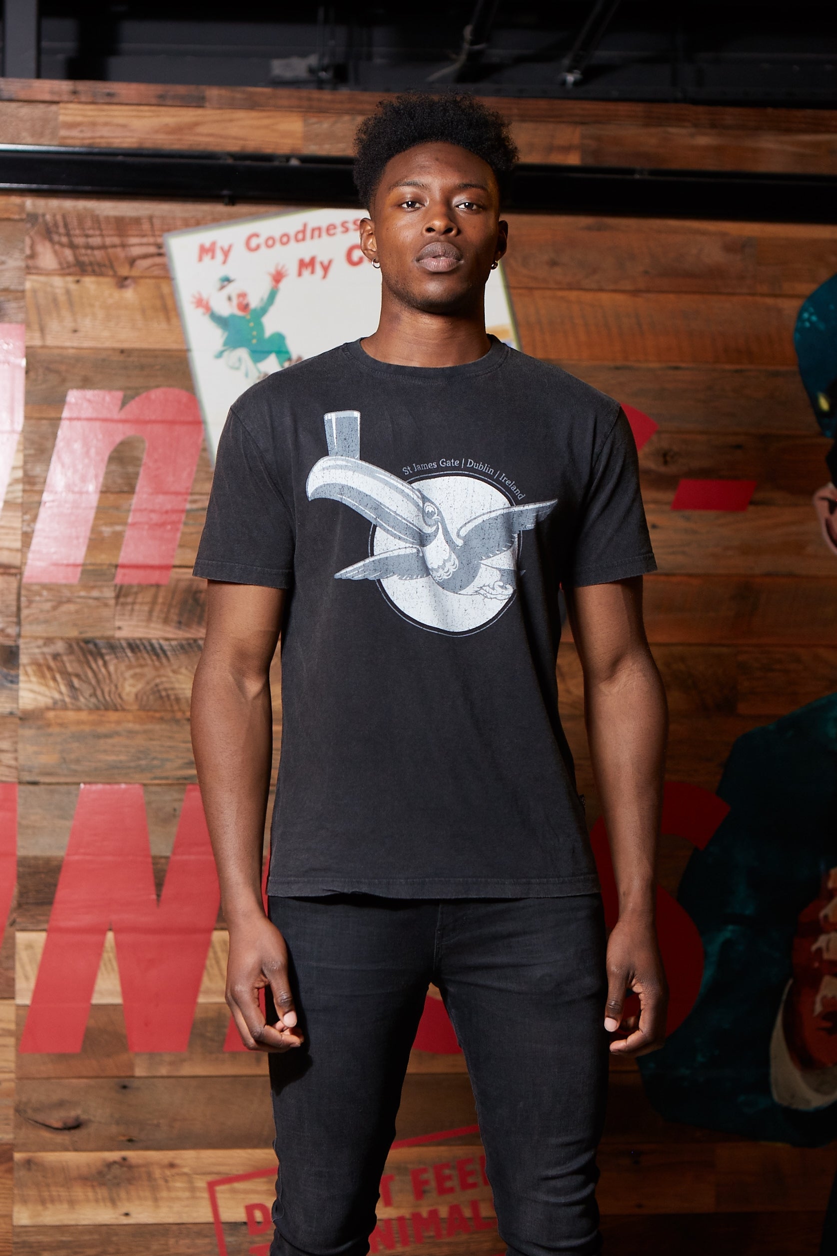 A man wearing a Guinness Premium Tee with Vintage Gilroy Toucan, a black t-shirt made of cotton fiber, standing next to a wooden wall.