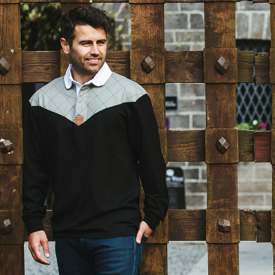 A man is standing next to a wooden fence wearing a Guinness Heritage Charcoal Grey & Black Long-Sleeve Rugby.