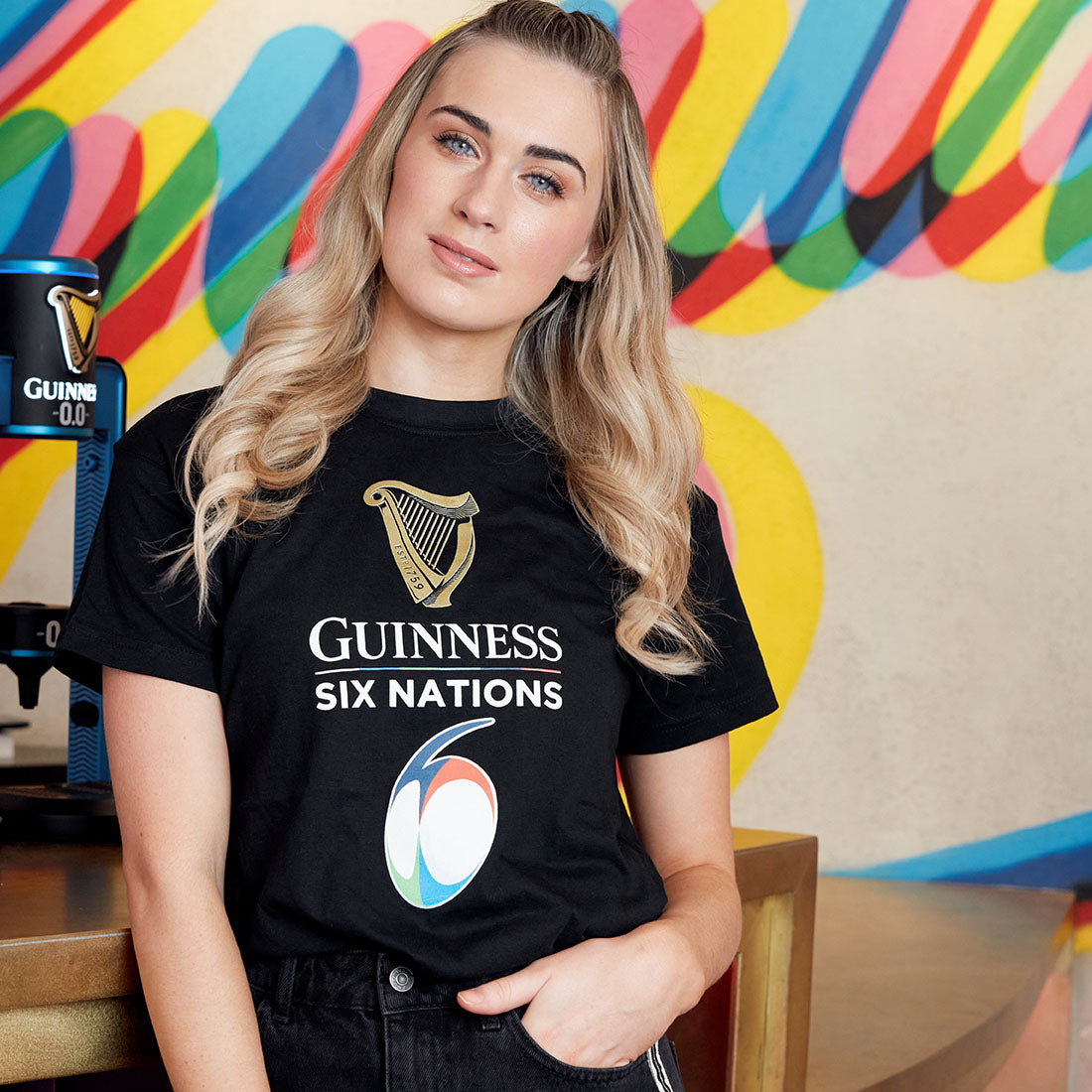 Woman in an official Guinness Six Nations Black Tee t-shirt standing in front of a colorful striped background.