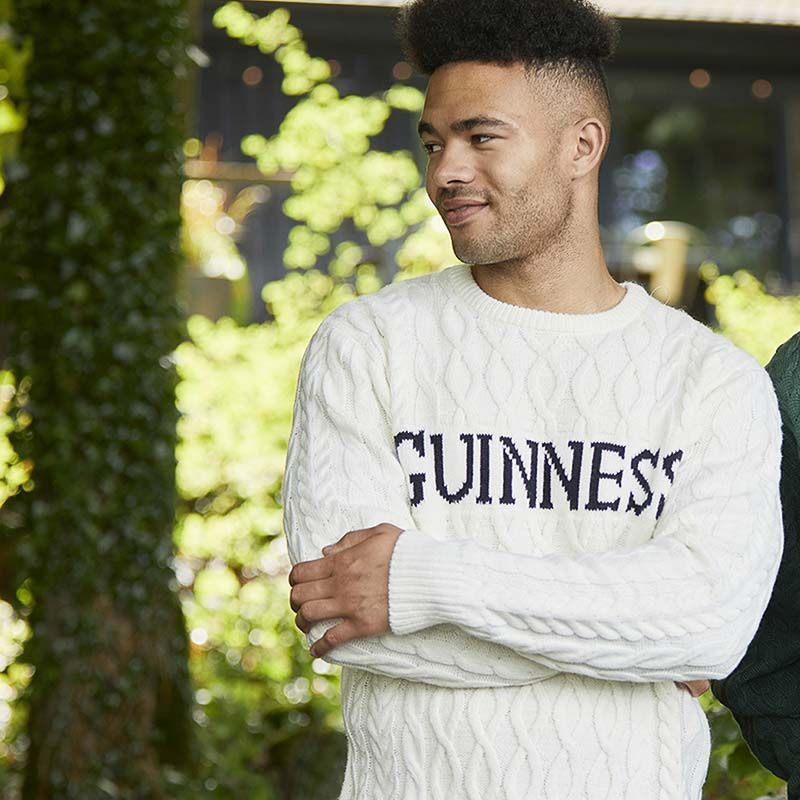 A person stands outdoors in classic Irish knitwear, wearing a Guinness Cream Cable Knit Jumper from Guinness Webstore US—a true winter essential with iconic style.