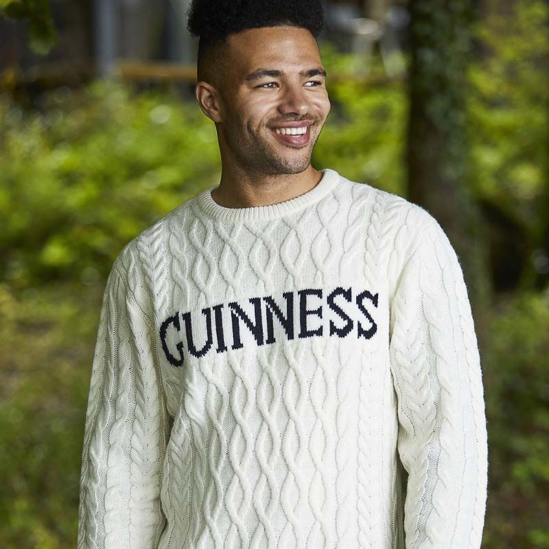 A man stands outdoors wearing a cream Guinness Cream Cable Knit Jumper from Guinness Webstore US, an Irish Aran winter essential with "GUINNESS" in bold black letters across the front.