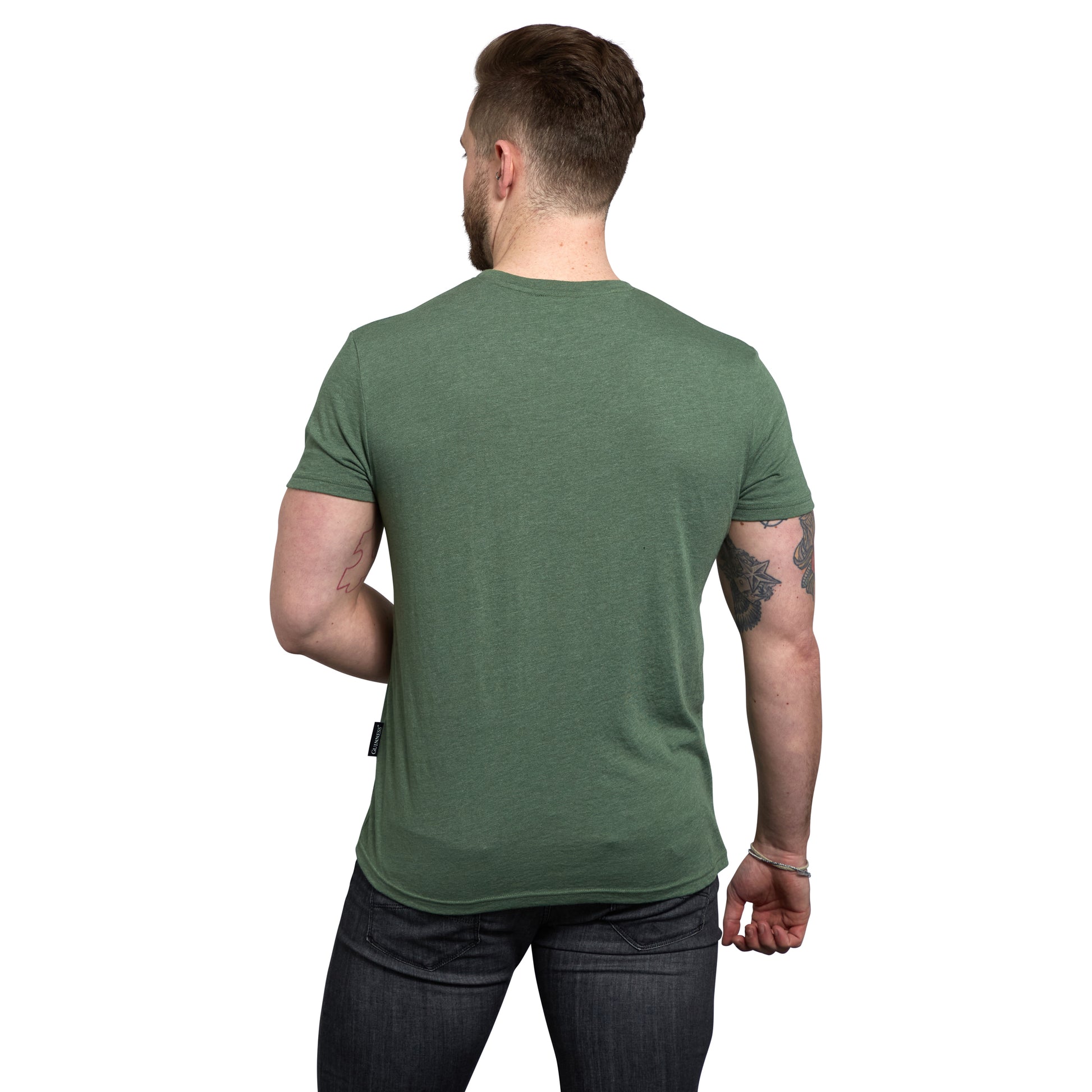 A man with short hair stands with his back to the camera in dark jeans, wearing the Guinness Green Distressed Label Tee crafted from a durable cotton blend.