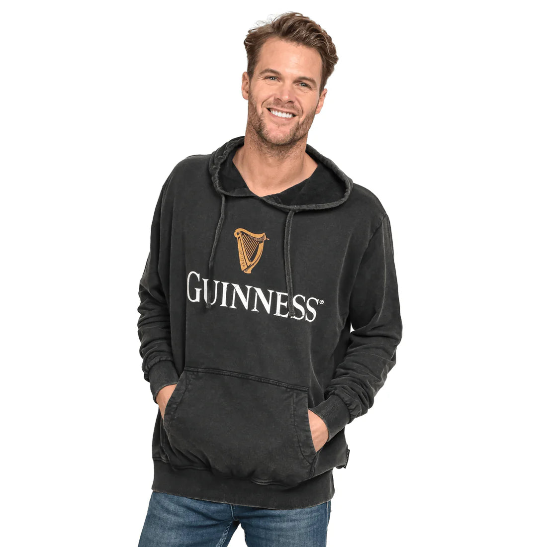 A man stands smiling while wearing a Guinness Premium Harp Hoodie from the Guinness Webstore US, featuring the iconic Guinness® Trademark Harp Logo on the front. He has his hands in the hoodie's front pocket and is dressed in blue jeans.