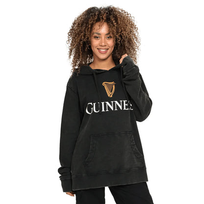 A person is wearing a black Guinness Premium Harp Hoodie from Guinness Webstore US, featuring a logo printed on the front, smiling while standing against a white background. The mid-weight brushed cotton jersey fabric ensures it is both comfortable and stylish.