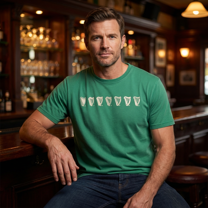 A man in a Guinness Green Evolution Harp Premium Tee featuring a white pint glass design sits at a wooden pub bar, looking at the camera.