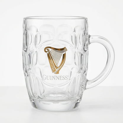 The Guinness Gold Harp Dimple Glass Mug – 20 oz Tankard from Guinness Webstore US features clear glass, a sturdy handle, and a gold harp with "Guinness" on the front—a classic Irish pub mug for your collection.