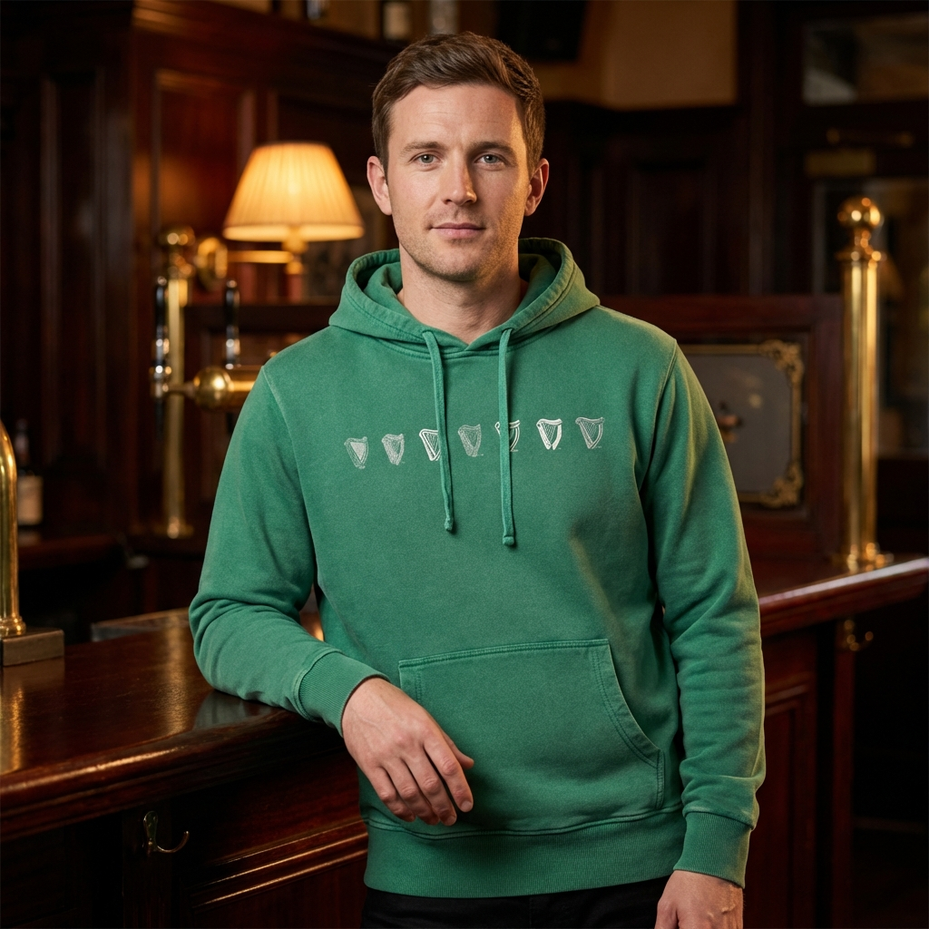 A man in a Guinness Evolution Harp Green Hoodie with white logos stands by a wooden bar in a warmly lit pub.