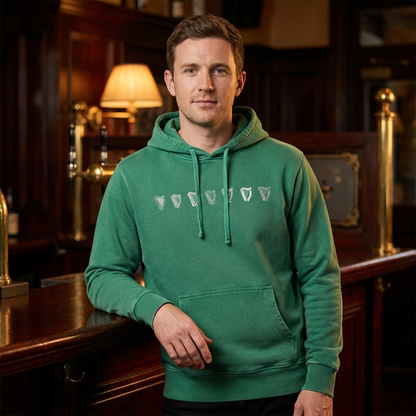 A man in a Guinness Evolution Harp Green Hoodie with white logos stands by a wooden bar in a warmly lit pub.