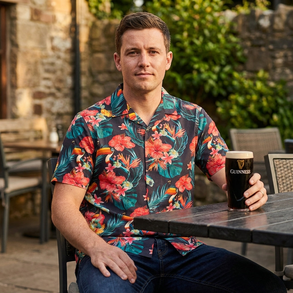 A man wearing a Guinness Toucan Hawaiian Shirt by Guinness sits at an outdoor table holding a pint of Guinness, with greenery and stone walls in the background.