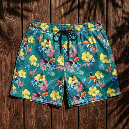 The Guinness Toucan Hawaiian Green Beach Shorts by Guinness Webstore US feature a tropical floral and toucan print, shown on a wooden surface dappled with leaf shadows.