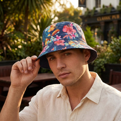 A man in a light shirt sits outdoors, touching the brim of his Guinness Hawaiian Reversible Bucket Hat from Guinness Webstore US, which features a colorful floral pattern.