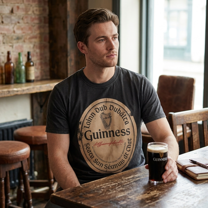A man sits at a wooden table in a pub holding a glass of Guinness, wearing the Guinness Black Distressed Label Tee by Guinness, with bottles and a window in the background.