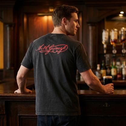 A man stands at a bar holding a pint of dark beer, wearing the Guinness Black Distressed Label Tee by Guinness, featuring red script text on the back, in a dimly lit setting.