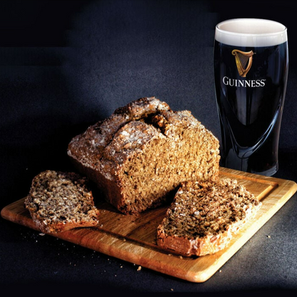 A loaf of brown bread partially sliced on a wooden board next to a full pint of Guinness beer, all set against a dark background, evokes the rich Guinness flavor found in our exclusive **Guinness Bread Kit** from **Guinness Webstore US** for home baking.