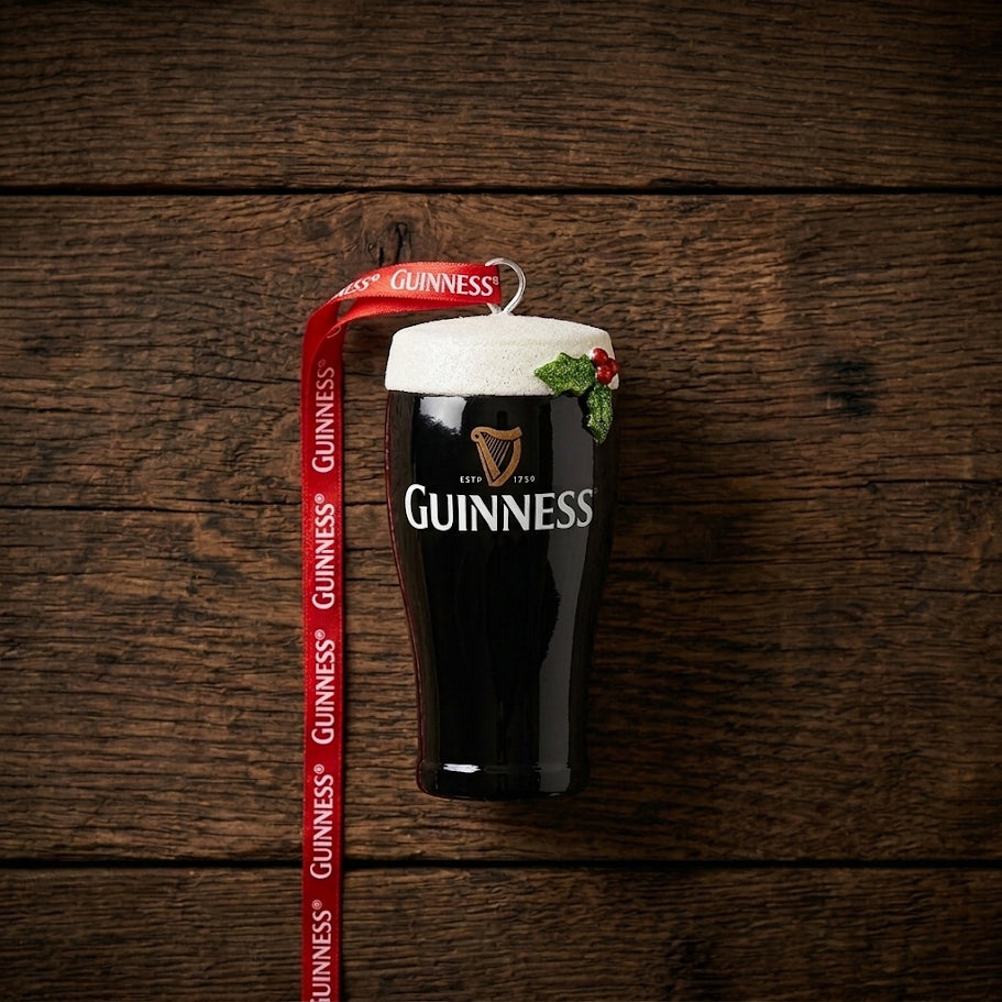 The Guinness Christmas Pint Decoration, featuring a pint glass shape with foam detail and holly, sits on a wooden surface and is attached to a red Guinness-branded ribbon—making it the perfect addition to your collection of Guinness Christmas decorations.