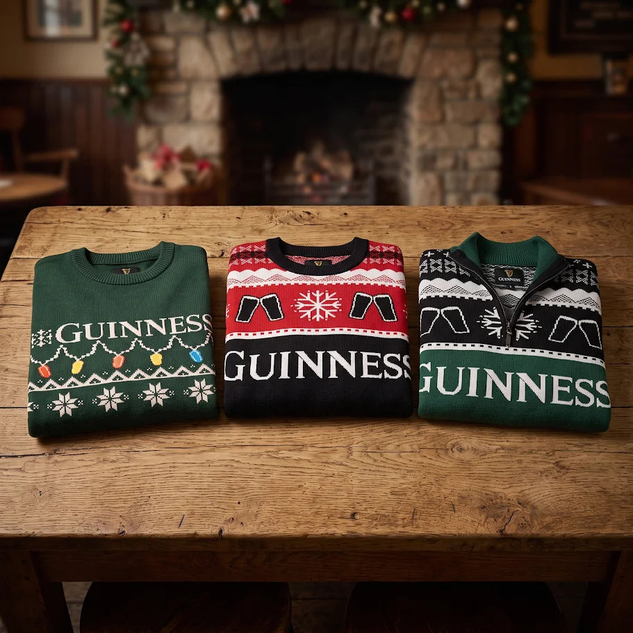 Three folded Guinness Holiday Quarter Zip Sweaters in green and black, featuring festive patterns, are displayed on a wooden table in front of a fireplace decorated for the holidays.