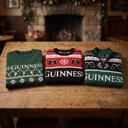 Three folded Guinness Holiday Quarter Zip Sweaters in green and black, featuring festive patterns, are displayed on a wooden table in front of a fireplace decorated for the holidays.