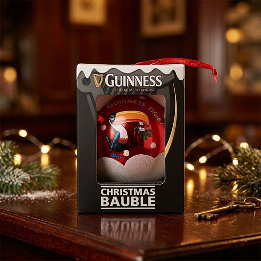 A Guinness Toucan Glass Bauble by Guinness, elegantly boxed, rests on a wooden table surrounded by pine branches and keys in a warmly lit room—creating the perfect festive touch for any Guinness Christmas tree decoration collection.