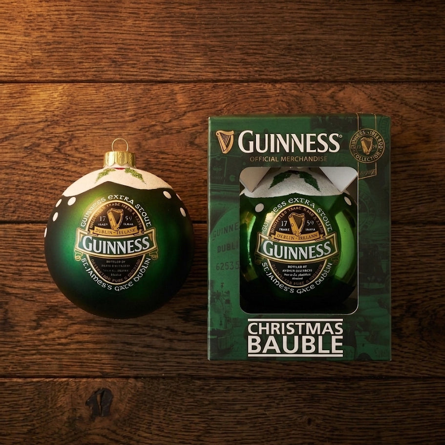 A festive Guinness Green Collection Gloss Bauble sits beside its packaging on a wooden surface. This Christmas ornament is designed to resemble a classic Guinness bottle cap and label, featuring the iconic green, black, and gold colors in tribute to Guinness Ireland.