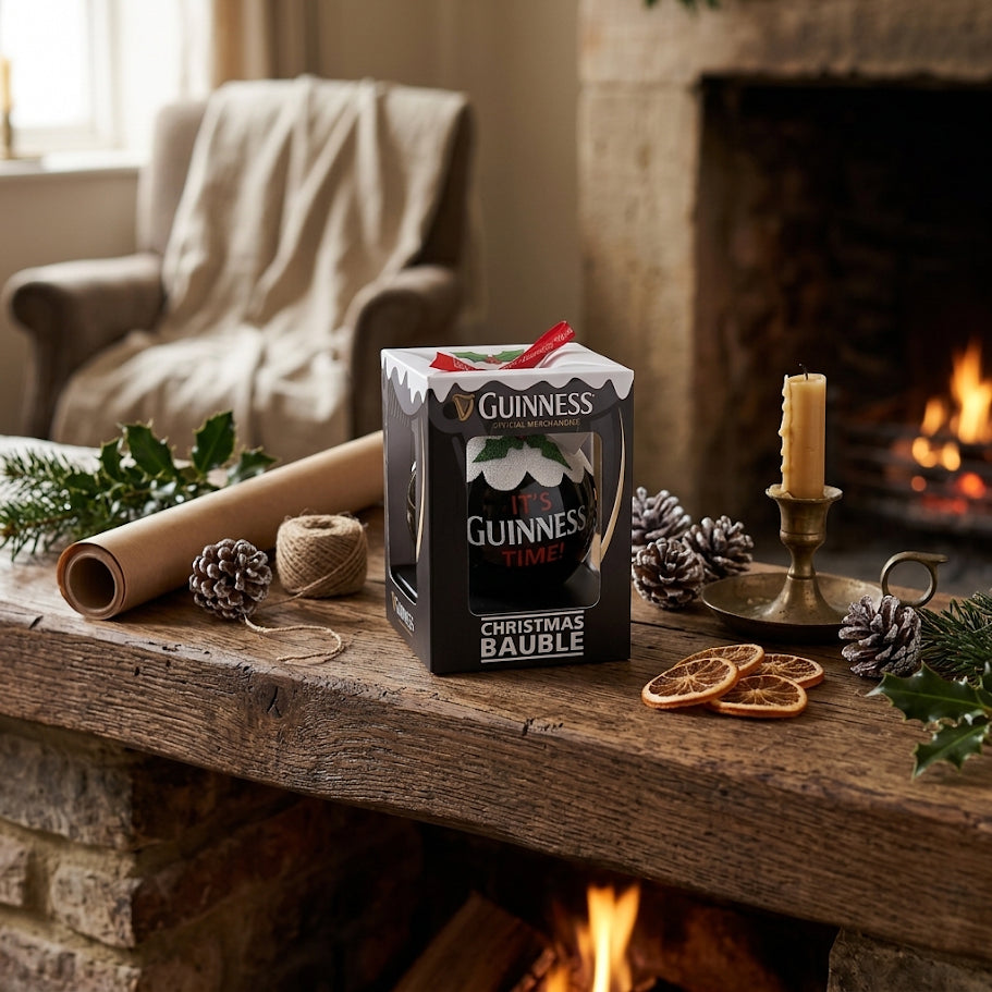 A Guinness Christmas Holiday Pudding Bauble in a box sits on a rustic mantel decorated with pinecones, dried oranges, a candle, and greenery near a lit fireplace.