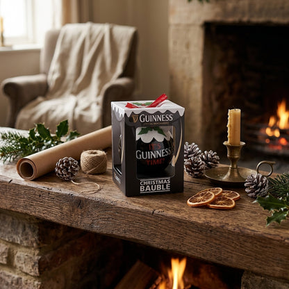 A Guinness Christmas Holiday Pudding Bauble in a box sits on a rustic mantel decorated with pinecones, dried oranges, a candle, and greenery near a lit fireplace.