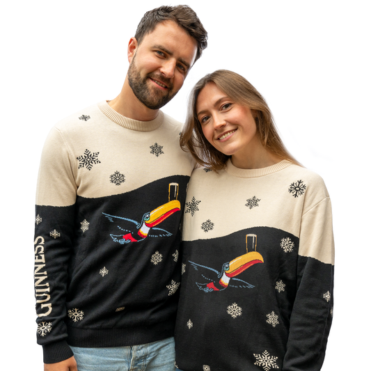 A smiling couple wearing matching Guinness Holiday Black and Cream Toucan Sweaters from the Guinness Webstore US, featuring a festive style with a snowflake design and "GUINNESS" emblazoned on their sleeves, pose proudly against a plain white background.